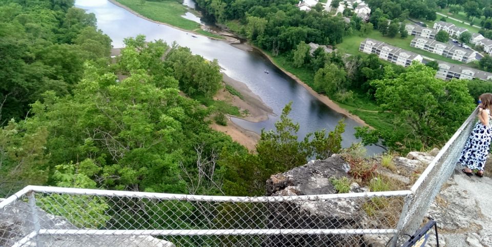 View from a cliff overlooking a river, with trees and houses on the far bank. A person stands by a fence on the right