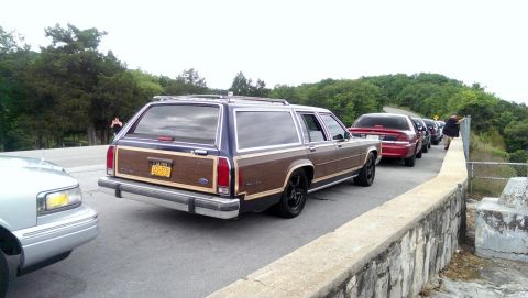 2000 Mercury Grand Marquis - A line of cars, including a wood-paneled station wagon, are parked on a road next to a stone wall.