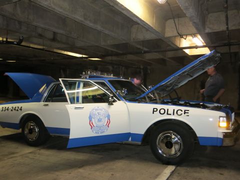 1991 Ford LTD Crown Victoria - blue-and-white police car lettered POLICE with raised hood sits in a concrete garage as two men work on it.