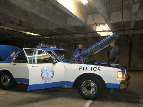1991 Ford LTD Crown Victoria - Two men inspect the engine of a blue-and-white POLICE car with its hood raised inside a concrete parking garage.
