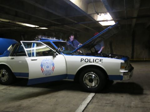 1991 Ford LTD Crown Victoria - white and blue police-marked sedan sits with its hood raised in a concrete parking garage while a man works on the engine.