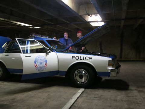 1991 Ford LTD Crown Victoria - Two men stand beside a white and blue police-lettered sedan with its hood up inside a dim concrete parking garage.