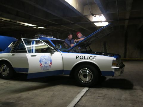 1991 Ford LTD Crown Victoria - white and blue sedan marked POLICE has its hood raised while two men work on it in a concrete parking garage.