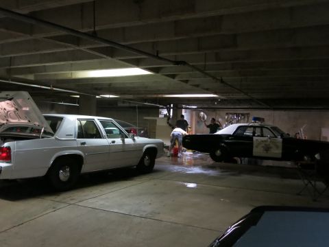 1991 Ford LTD Crown Victoria - white sedan with open trunk and a black highway-patrol-marked car sit in a parking garage as people wash and detail vehicles.