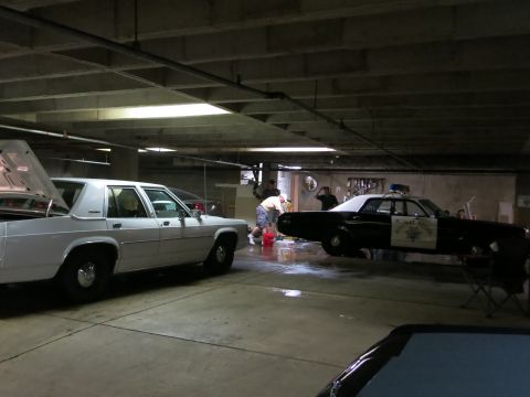 1991 Ford LTD Crown Victoria - white sedan and a black highway-patrol-style car are parked in a concrete garage while several people wash and prep the vehicles.