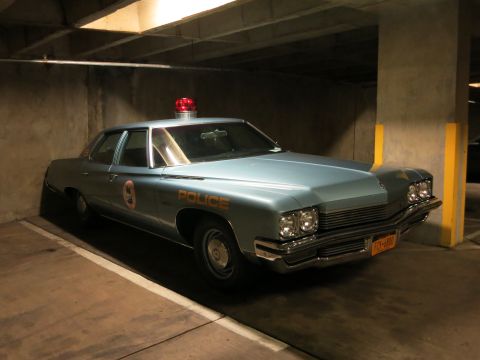 1991 Ford LTD Crown Victoria - light blue four-door police sedan with a single red roof beacon and door number 9 is parked in a concrete parking garage.