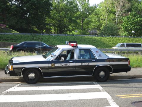 1991 Ford LTD Crown Victoria - black and white Trumbull Police sedan with roof lightbar is parked at a roadside crosswalk as cars pass on a tree-lined highway.