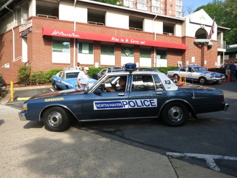 1991 Ford LTD Crown Victoria - blue North Haven Police sedan with roof lightbar and unit 13 is parked outside a brick pizza-and-pasta restaurant near other classic cars.