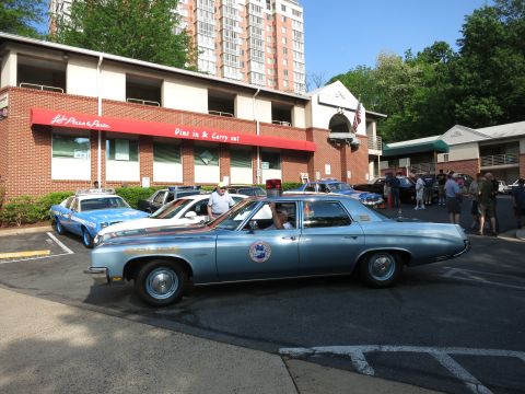 1991 Ford LTD Crown Victoria - light blue vintage police-marked sedan sits with other classic cars in a lot outside a brick restaurant as a crowd gathers.