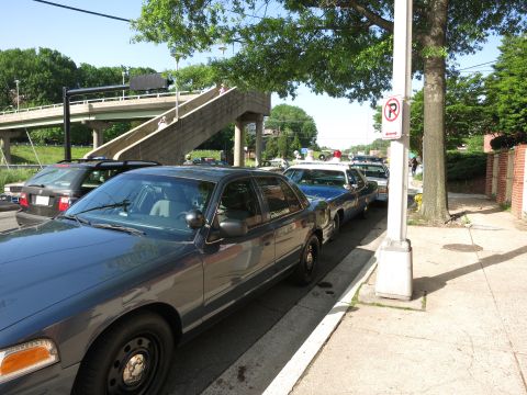 1991 Ford LTD Crown Victoria - Several dark sedans, some with roof lights, are parked nose to tail along a curb beside a pedestrian overpass and no-parking sign.