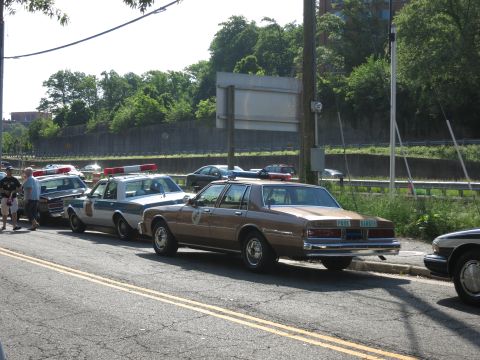 1991 Ford LTD Crown Victoria - line of vintage police sedans, including a brown Park Police car, is parked along a curb beside a highway guardrail.