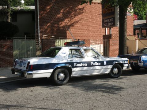 1991 Ford LTD Crown Victoria - blue and white Boston Police sedan with roof lightbar is parked on a street in front of a brick Best Western hotel sign.