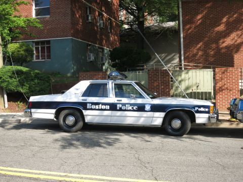 1991 Ford LTD Crown Victoria - blue and white Boston Police sedan with roof lightbar is parked along a curb beside brick apartment buildings.