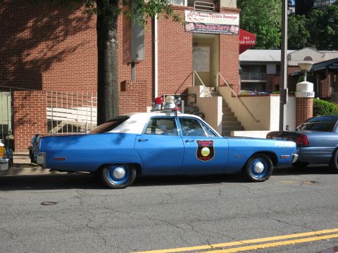1991 Ford LTD Crown Victoria - blue and white vintage police sedan with a shield door emblem and roof beacon is parked on a street beside a brick building.