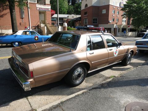 1991 Ford LTD Crown Victoria - brown four-door police sedan with roof lightbar and door emblem is parked on a street among other classic police cars near brick buildings.