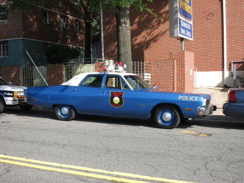 1991 Ford LTD Crown Victoria - blue and white sedan lettered POLICE with a shield emblem and dual roof beacons is parked along a curb by a brick building.