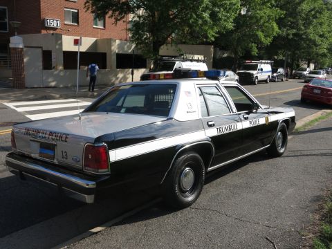 1991 Ford LTD Crown Victoria - black-and-white Ford LTD Crown Victoria marked Trumbull Police, unit 13, parked on a tree-lined city street with other patrol cars ahead.
