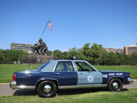 1991 Ford LTD Crown Victoria - blue-and-gray Massachusetts State Police Ford sedan, unit 90E, parked on grass with the Iwo Jima Memorial and U.S. flag behind it.