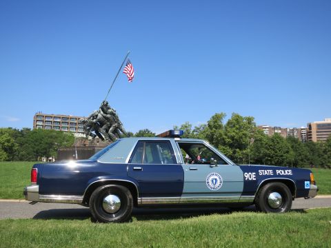 1991 Ford LTD Crown Victoria - Side profile of a blue-and-gray Massachusetts State Police Ford sedan, unit 90E, parked before the Iwo Jima Memorial under a clear sky.