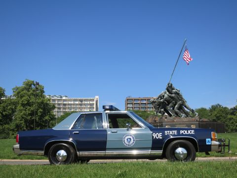 1991 Ford LTD Crown Victoria - blue-and-gray Massachusetts State Police Ford sedan, unit 90E, parked on a path with the Iwo Jima Memorial and flag in the background.