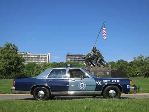 1991 Ford LTD Crown Victoria - blue-and-gray Massachusetts State Police Ford sedan, unit 90E, sits on grass with the Iwo Jima Memorial centered behind it.