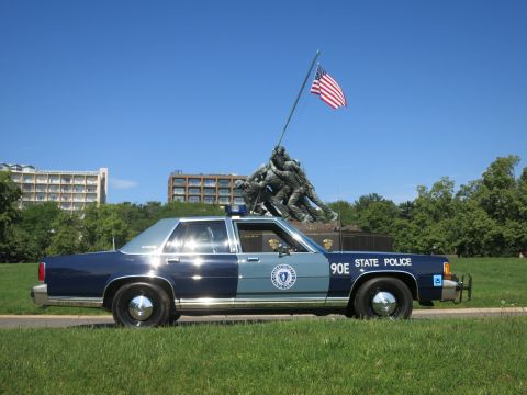 1991 Ford LTD Crown Victoria - blue-and-gray Massachusetts State Police Ford sedan, unit 90E, parked on grass with the Iwo Jima Memorial and flag behind it.