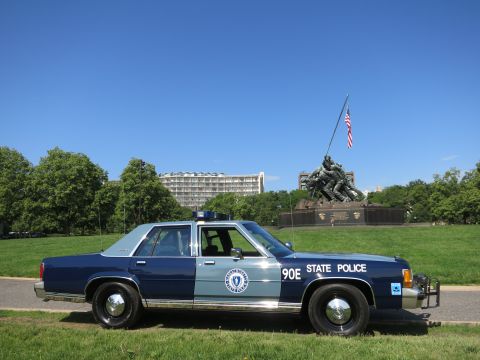 1991 Ford LTD Crown Victoria - Side profile of a blue-and-gray Massachusetts State Police Ford sedan, unit 90E, parked on grass before the distant Iwo Jima Memorial.