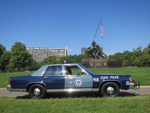 1991 Ford LTD Crown Victoria - blue-and-gray Massachusetts State Police Ford sedan, unit 90E, with a push bar, parked on grass before the Iwo Jima Memorial.