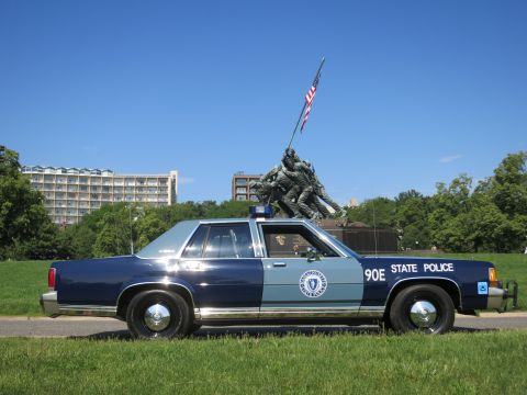 1991 Ford LTD Crown Victoria - blue-and-gray Massachusetts State Police Ford sedan, unit 90E, parked on grass with the Iwo Jima Memorial and flag behind.