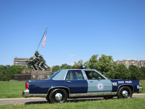1991 Ford LTD Crown Victoria - Rear-quarter view of a blue-and-gray Massachusetts State Police Ford sedan, unit 90E, parked on grass before the Iwo Jima Memorial.