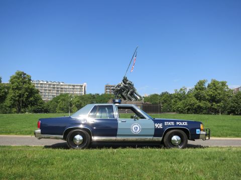 1991 Ford LTD Crown Victoria - two-tone blue Massachusetts State Police cruiser marked 90E sits on a roadside lawn with the Iwo Jima Marine Corps Memorial behind it.