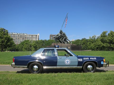 1991 Ford LTD Crown Victoria - Rear-quarter view of a navy and light-blue Massachusetts State Police sedan, with the flag-raising Marine memorial and a building in the ba…