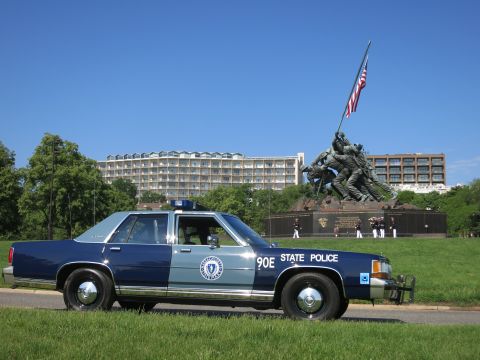 1991 Ford LTD Crown Victoria - Side view of a Massachusetts State Police cruiser marked 90E with the Marine Corps War Memorial and a row of uniformed people behind it.
