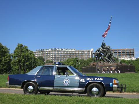 1991 Ford LTD Crown Victoria - two-tone blue State Police sedan sits on a lawn in front of the Marine Corps flag-raising memorial under a clear sky.