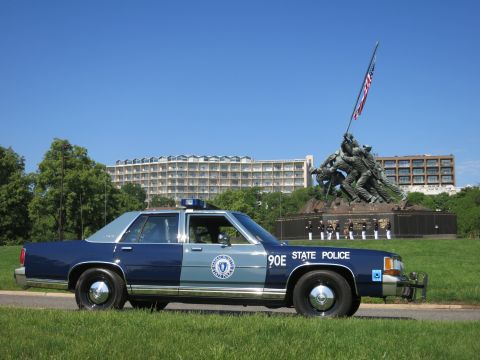 1991 Ford LTD Crown Victoria - Profile of a navy and pale-blue Massachusetts State Police sedan with the Marine Corps War Memorial statue rising behind it.