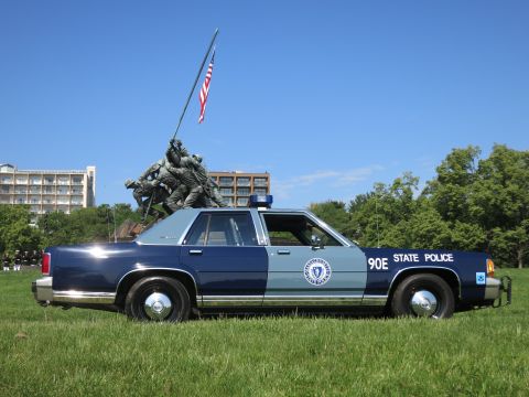 1991 Ford LTD Crown Victoria - navy and light-blue Massachusetts State Police sedan on grass with the Marine Corps flag-raising memorial behind it.