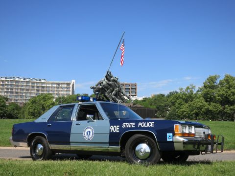 1991 Ford LTD Crown Victoria - Front three-quarter view of a two-tone blue State Police cruiser with a brush guard, parked before the Marine memorial.