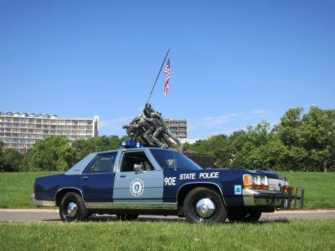 1991 Ford LTD Crown Victoria - darker navy two-tone State Police sedan with push bar sits below the Marine Corps flag-raising memorial.