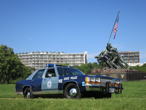 1991 Ford LTD Crown Victoria - two-tone blue State Police sedan with a front license plate and brush guard sits before the Marine memorial.