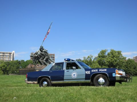 1991 Ford LTD Crown Victoria - Profile of a navy and pale-blue State Police cruiser on grass with the Marine Corps flag-raising memorial behind.
