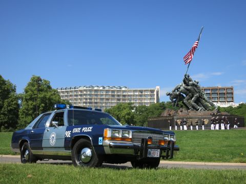 1991 Ford LTD Crown Victoria - two-tone blue State Police sedan with brush guard before the Marine memorial, rows of uniformed personnel lined up behind.