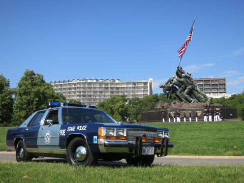 1991 Ford LTD Crown Victoria - navy and light-blue State Police cruiser before the Marine Corps War Memorial with uniformed personnel arrayed behind.
