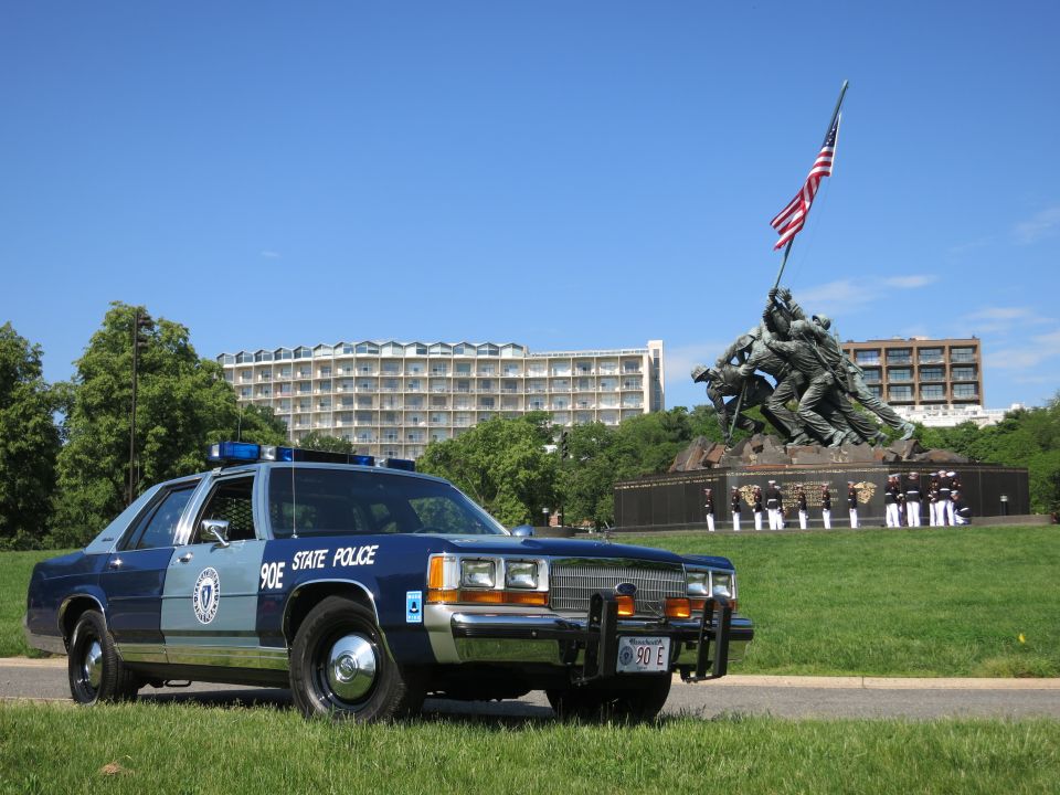 Front three-quarter of blue Massachusetts State Police 90E sedan with uniformed personnel near Iwo Jima Memorial