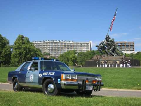 1991 Ford LTD Crown Victoria - two-tone blue State Police sedan with front plate and brush guard, rows of uniformed personnel behind near the Marine memorial.