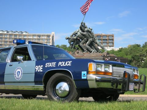 1991 Ford LTD Crown Victoria - Close front three-quarter of a navy and light-blue State Police cruiser marked 90E directly below the Marine flag-raising memorial.