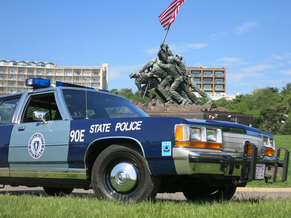 Close front three-quarter of blue Massachusetts State Police 90E sedan beneath Iwo Jima Memorial