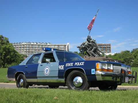 1991 Ford LTD Crown Victoria - two-tone blue Massachusetts State Police cruiser marked 90E sits on grass with the Iwo Jima Memorial and an American flag behind it.
