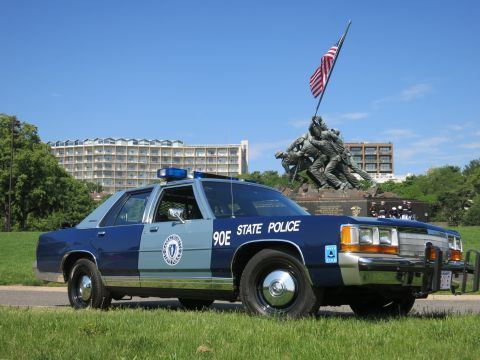 1991 Ford LTD Crown Victoria - blue and light-blue Massachusetts State Police sedan marked 90E parked on grass with the Iwo Jima Memorial in the background.