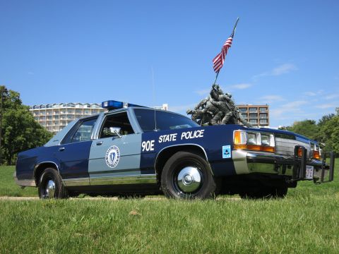 1991 Ford LTD Crown Victoria - Front three-quarter view of a blue Massachusetts State Police sedan with a push bar, parked on grass before the Iwo Jima Memorial.