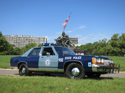 1991 Ford LTD Crown Victoria - Side profile of a blue and light-blue Massachusetts State Police sedan marked 90E on grass with the Iwo Jima Memorial in view.